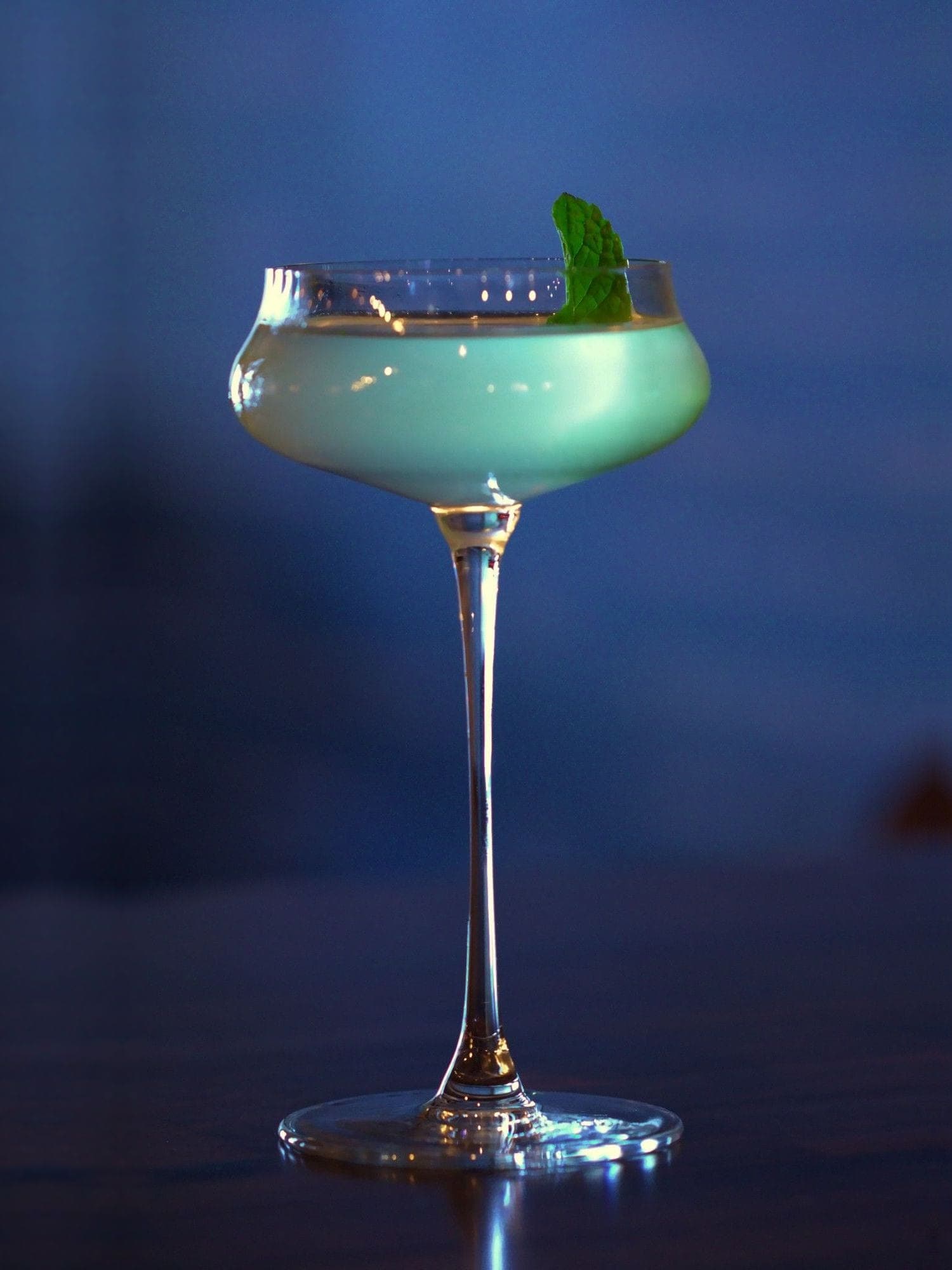 Luckeys London Dry, mint, lime and cucumber — pale green in a tall-stemmed coupe with a mint leaf, against the blue of the bar at dusk